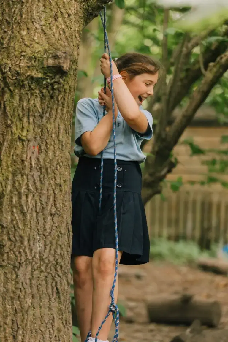 Girl climbing on a rope in the outdoor playground of St Katharines Primary School, surrounded by trees, fostering outdoor activities and physical development.